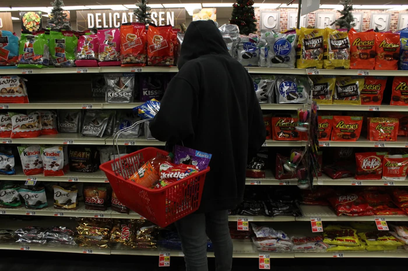 A hooded figure with a red shopping basket standing closed-off in front of shelves of candy