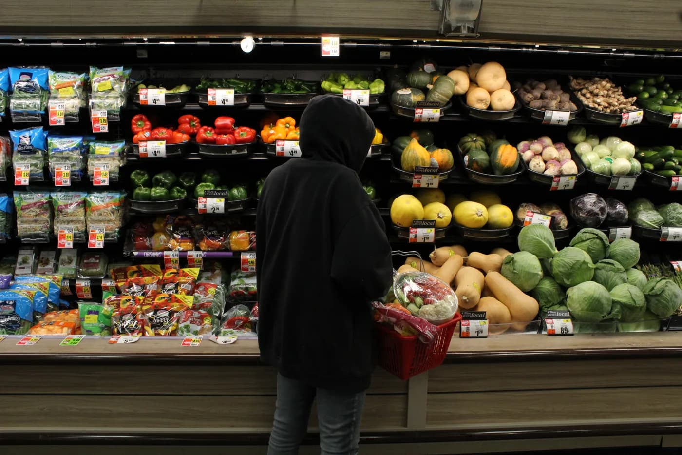 The hooded figure with an open, thoughtful posture choosing vegetables in a brightly lit produce aisle