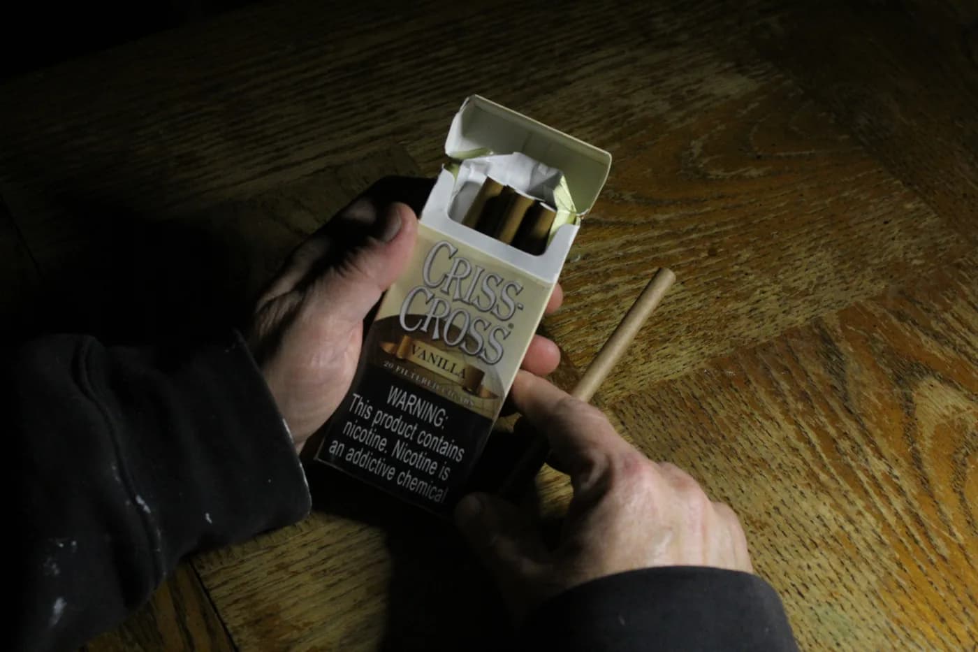 Hands holding an opened box of cigarettes over a wooden table in dark, underexposed light