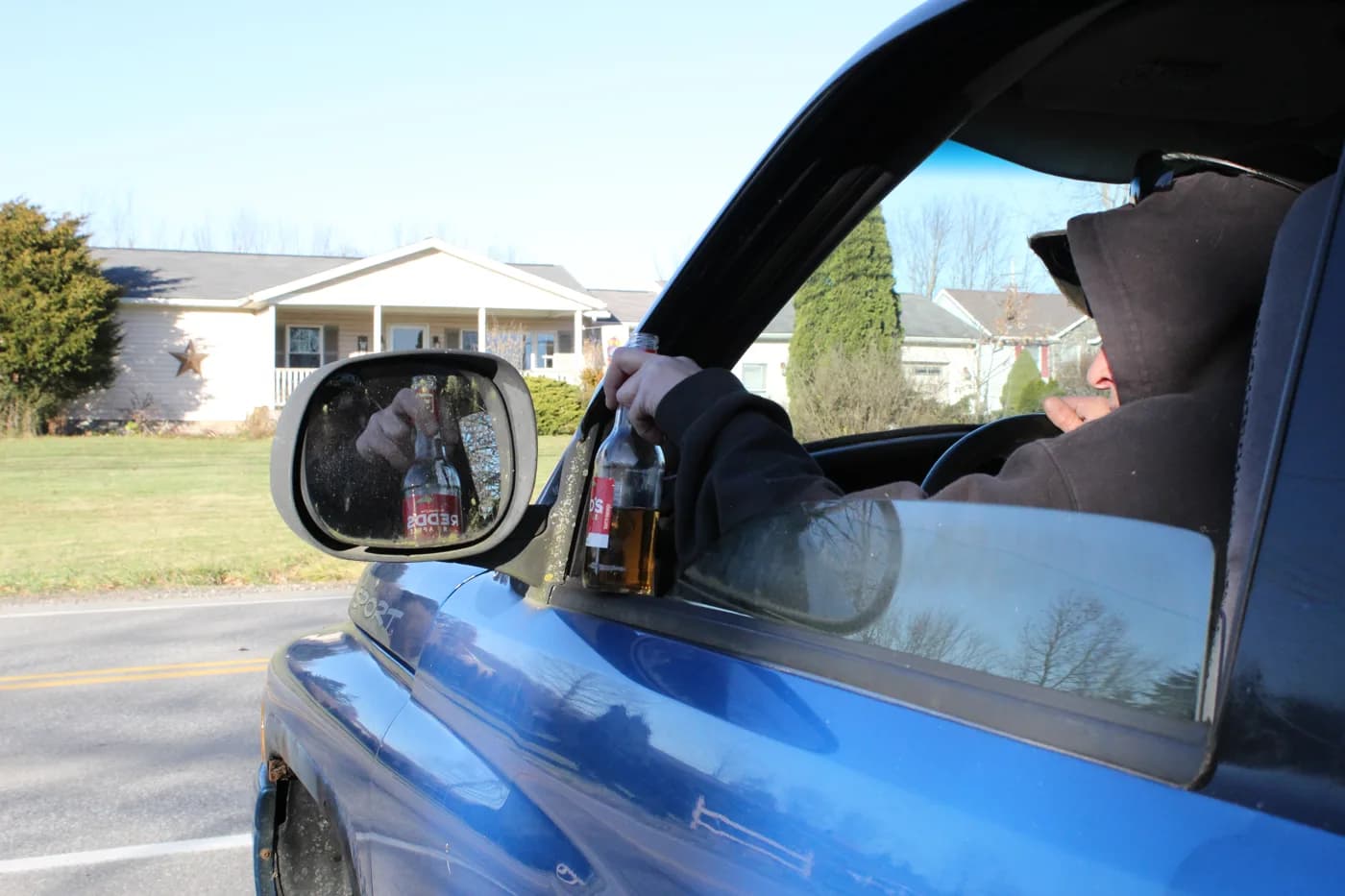 A hooded figure in a blue truck holding a bottle of alcohol out the window, the bottle reflected in the side mirror