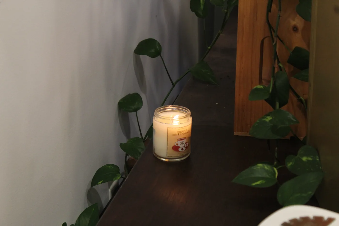 A small candle glowing on a dark shelf among synthetic vines against a white wall