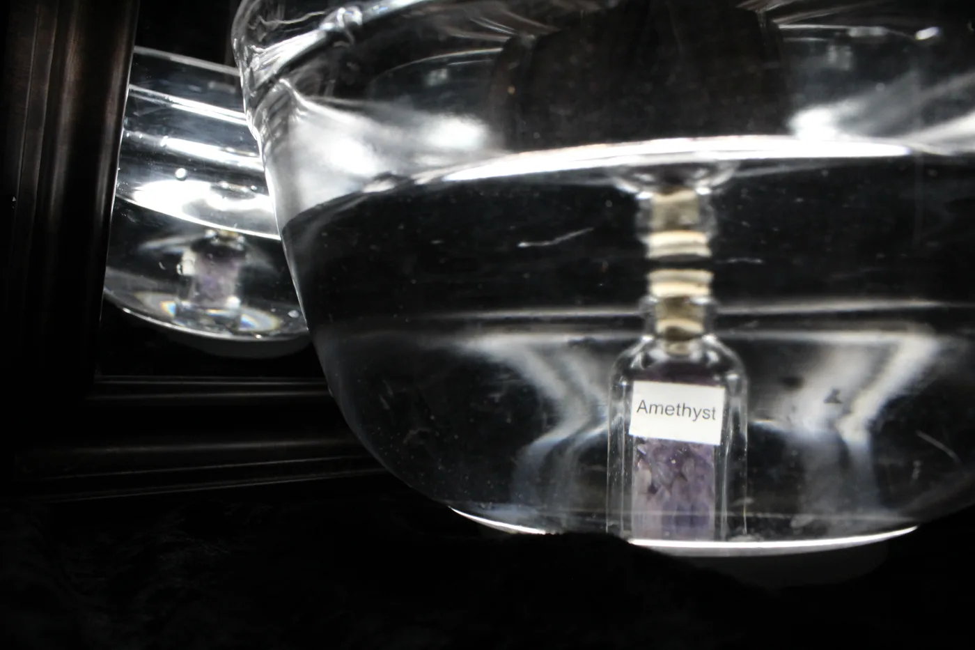 The amethyst bottle submerged in a bowl of water beside a mirror reflecting the scene