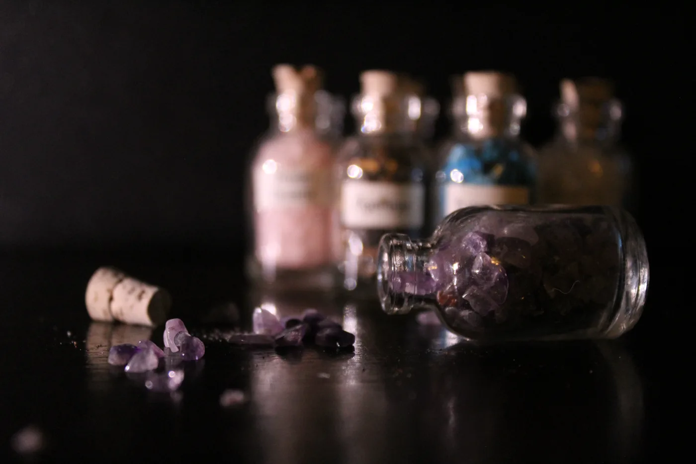 A small bottle tipped on its side with amethyst stones spilled across a dark reflective table, the other bottles blurred behind