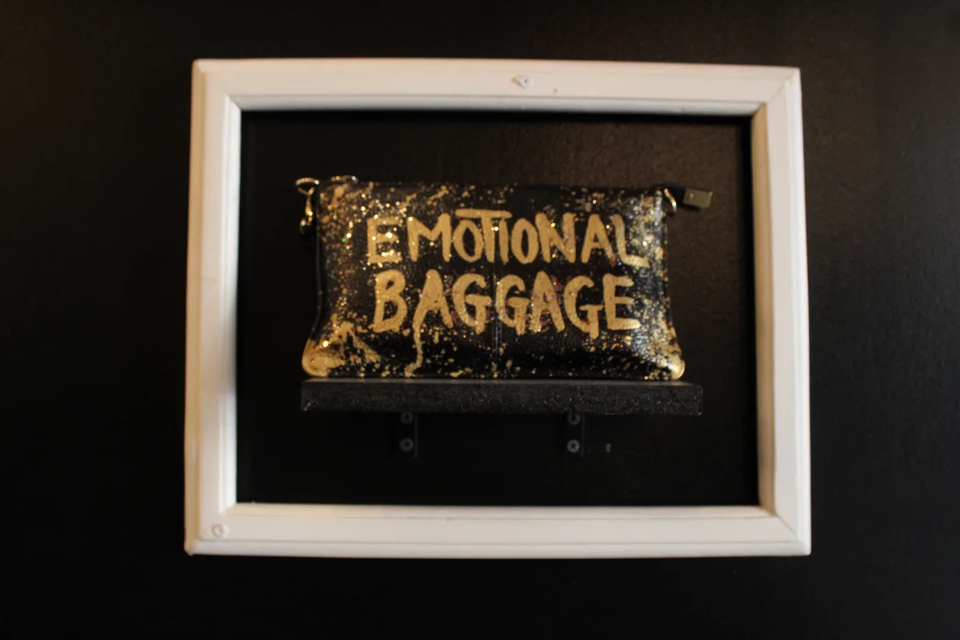 A black purse splattered with gold paint reading Emotional Baggage, displayed on a shelf inside a white picture frame