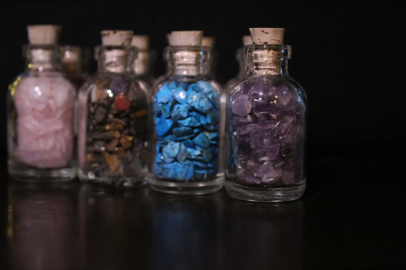 Four small corked bottles of colorful gemstones lined up against a dark background, the amethyst bottle in sharp focus